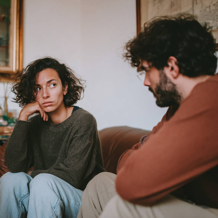 Couple on sofa in tense conversation, woman looking concerned with hand on chin as partner looks down, relationship conflict