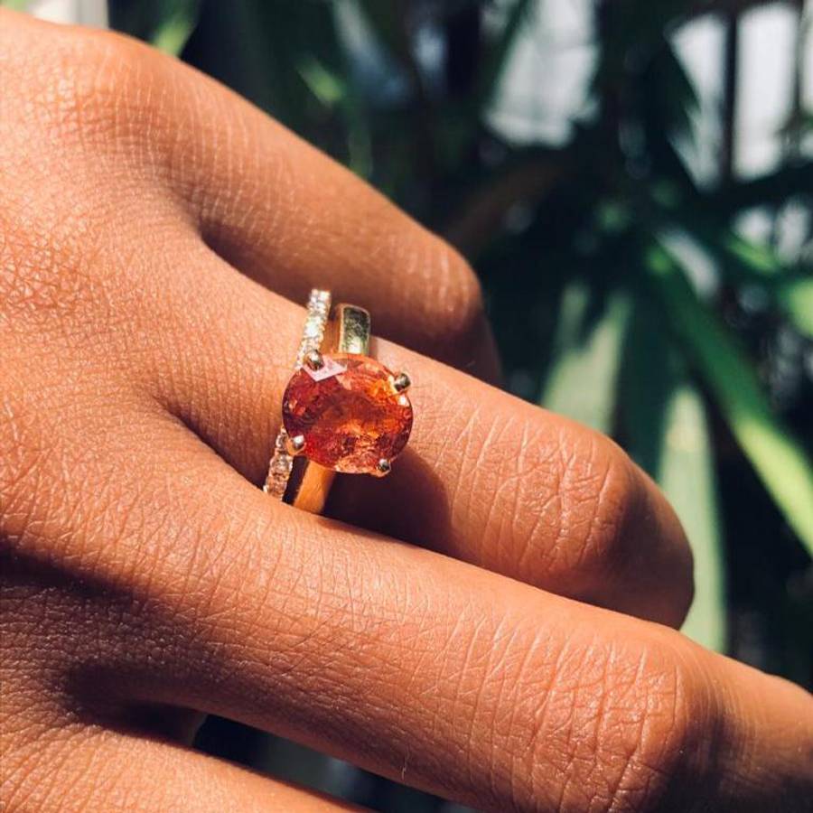 Close-up of a hand wearing a gold ring set with a vibrant round orange gemstone, paired with a slim diamond band, photographed in bright natural light with greenery in the background.