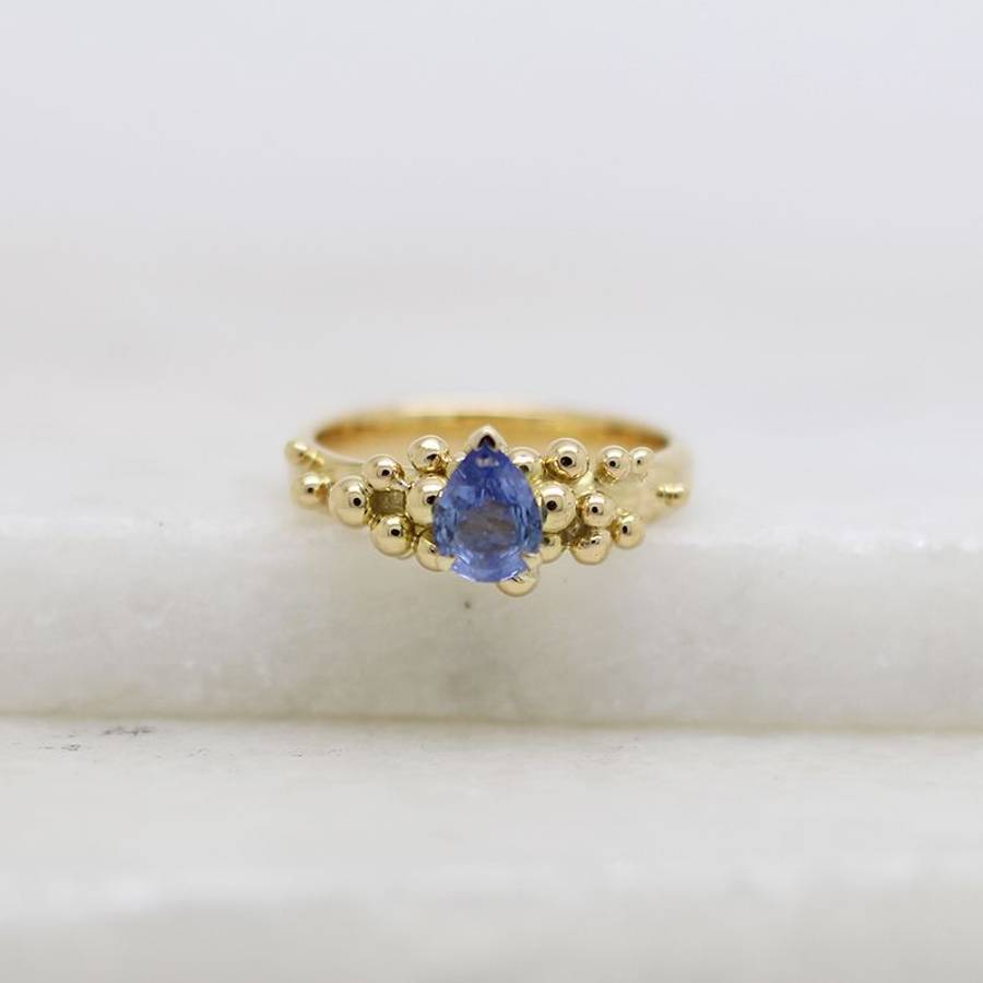 Gold ring with a pear-shaped blue gemstone centered among small gold bead accents, photographed on a white surface with a soft, neutral background.