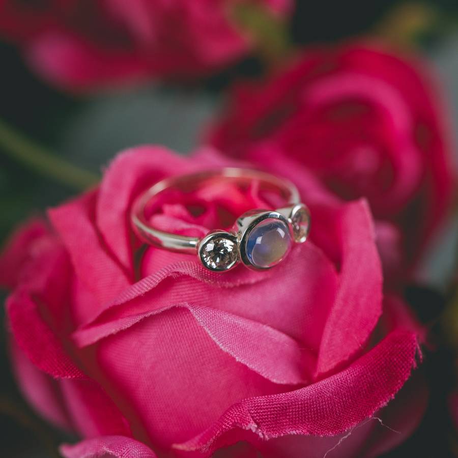 Silver ring with a central moonstone cabochon flanked by two clear gemstones, resting on bright pink rose petals with a softly blurred floral background.