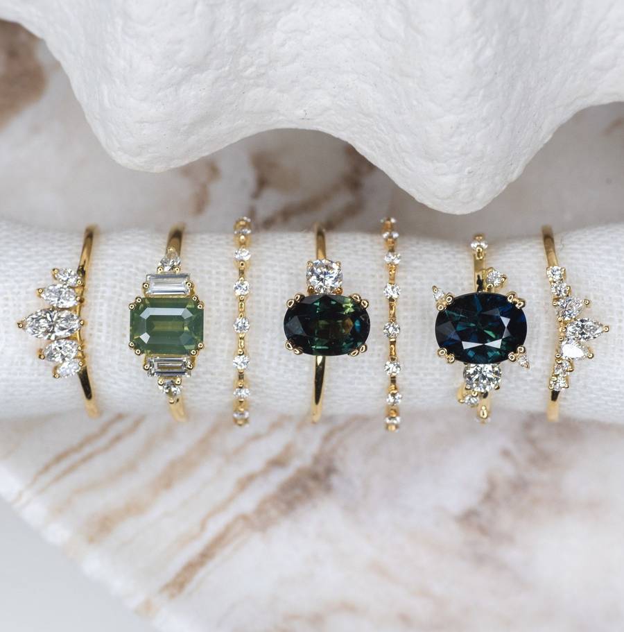 Row of delicate gold rings displayed on a white fabric roll, featuring a mix of diamond bands and gemstone rings with green and blue stones set in varied shapes and settings.