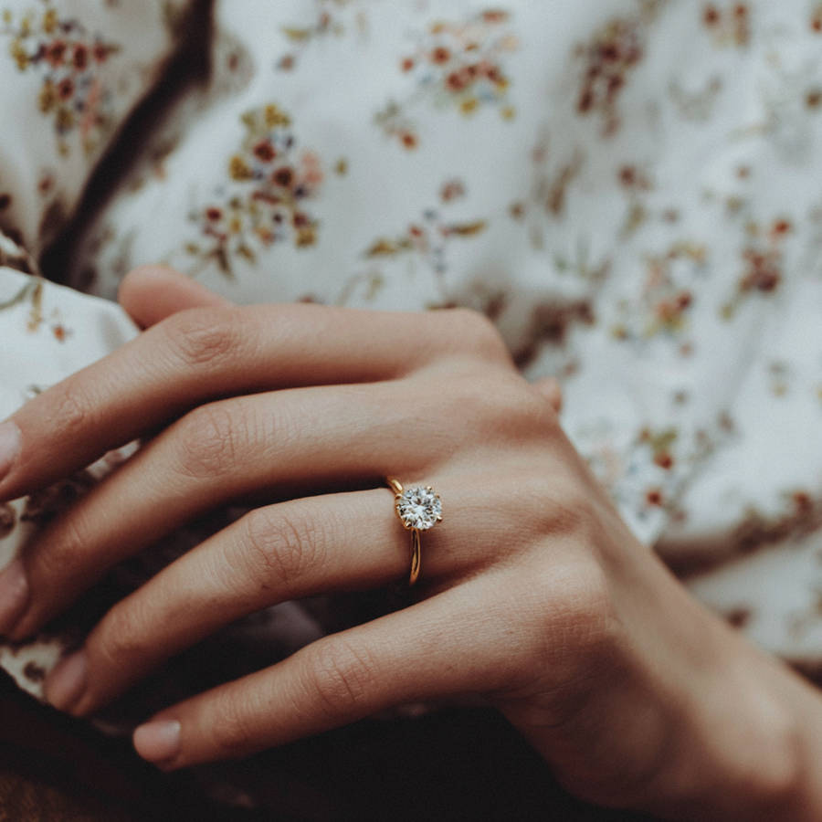 Close-up of a hand wearing a custom gold solitaire diamond engagement ring against a soft floral fabric background