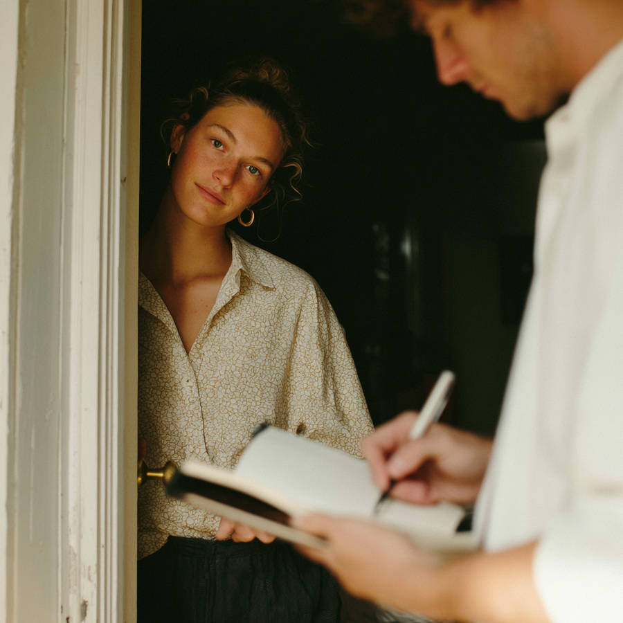 Woman leaning in a doorway smiling while a partner writes in a notebook during a quiet home moment
