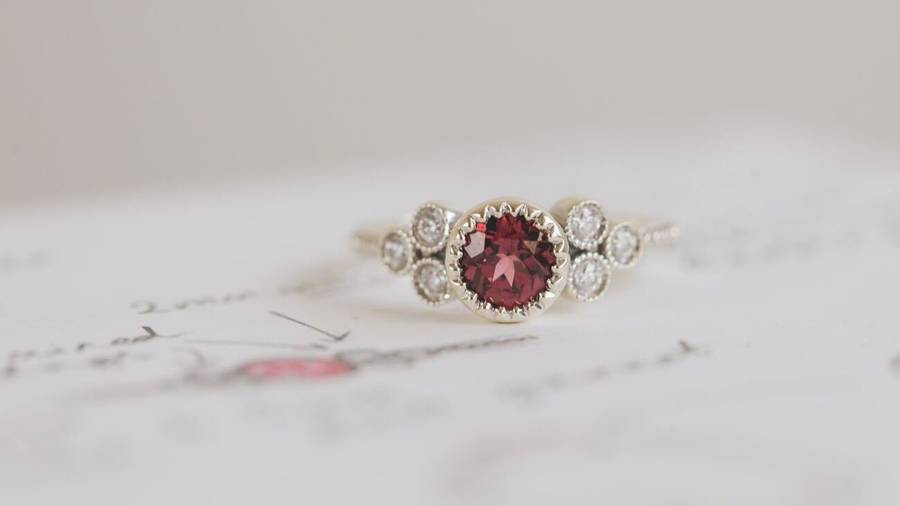 White gold ring with a round deep red garnet center stone, accented by clusters of small white diamonds on each side, displayed on a softly blurred sketch background.