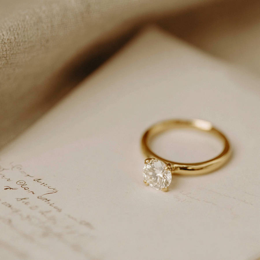 Gold solitaire engagement ring with a round diamond resting on an open sketchbook page, softly lit with a neutral background.