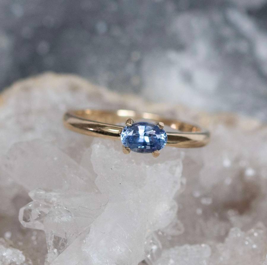 Yellow gold ring with an oval blue gemstone set in four prongs, resting on clear quartz crystals against a soft grey background