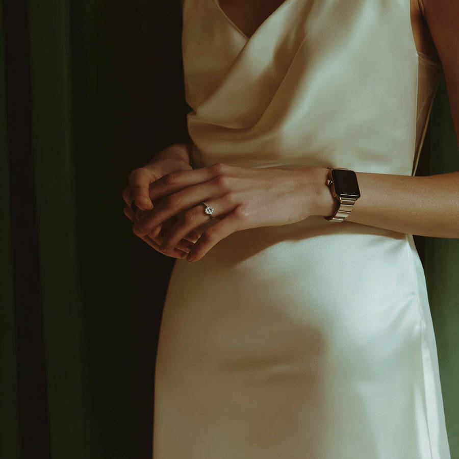 Close-up of woman in satin dress wearing a bespoke diamond engagement ring and gold smartwatch in soft light