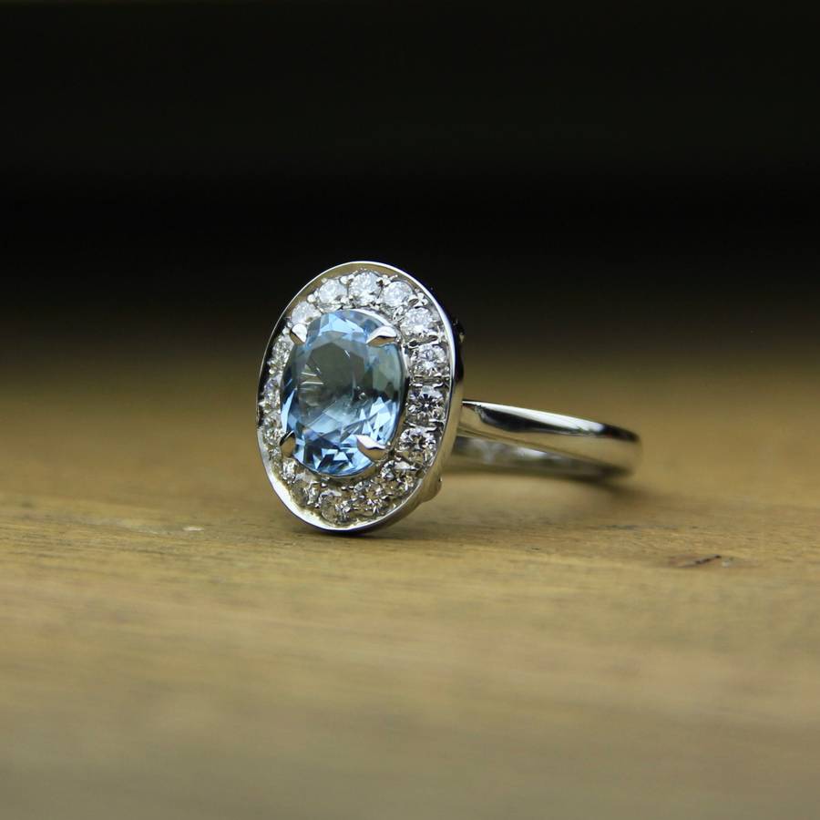 Oval light blue aquamarine ring with a diamond halo set in white metal, resting on a wooden surface with a softly blurred dark background