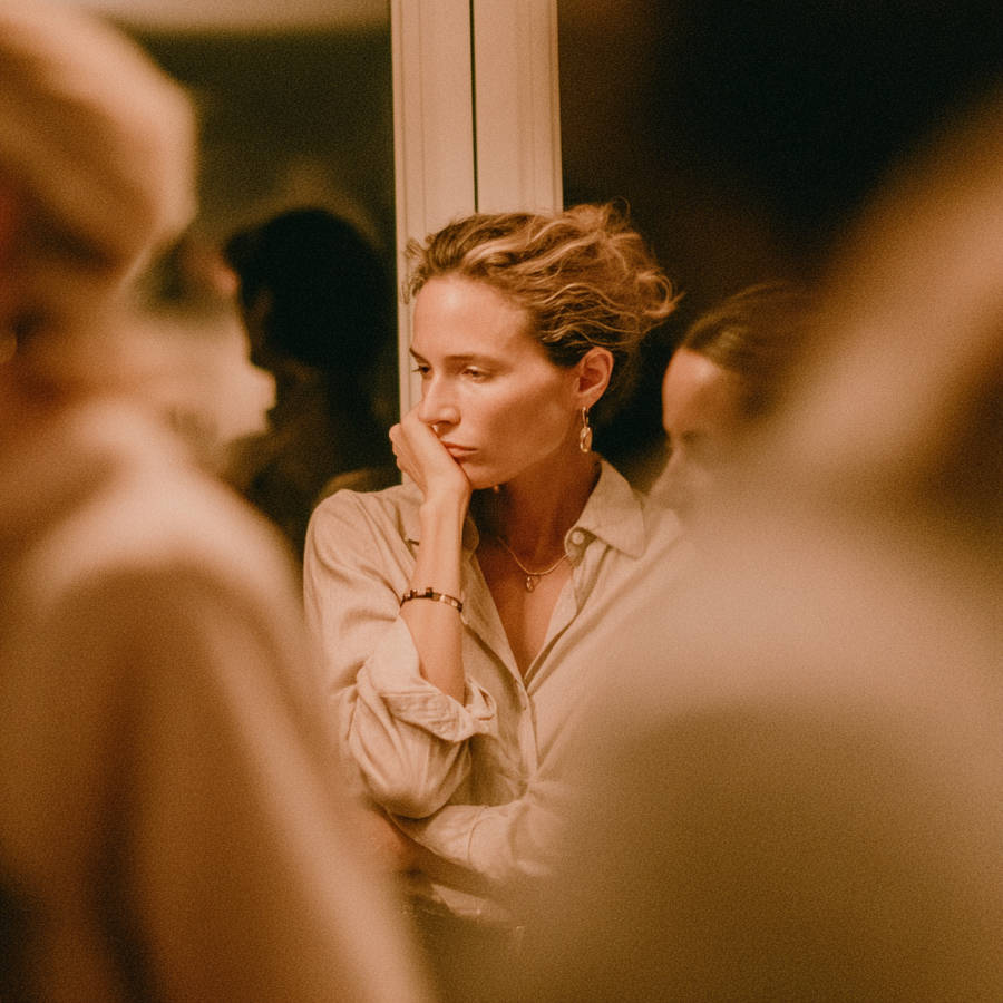 Pensive woman at a house party resting chin on hand, looking worried amid blurred guests in warm indoor lighting
