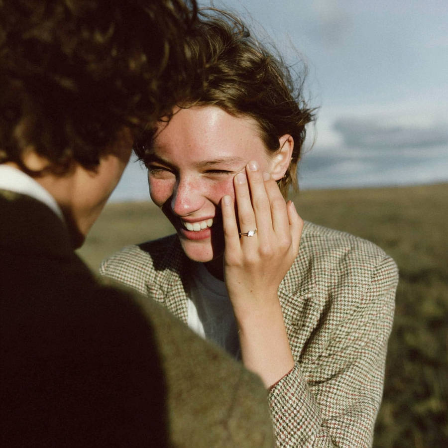 Smiling woman shows her custom engagement ring on her hand, laughing with partner outdoors by the coast
