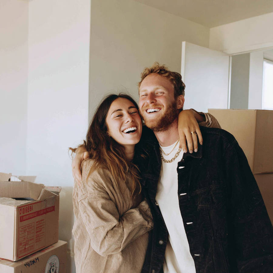 Happy couple hugging while moving into a new home, surrounded by cardboard boxes; woman’s ring visible on her hand.