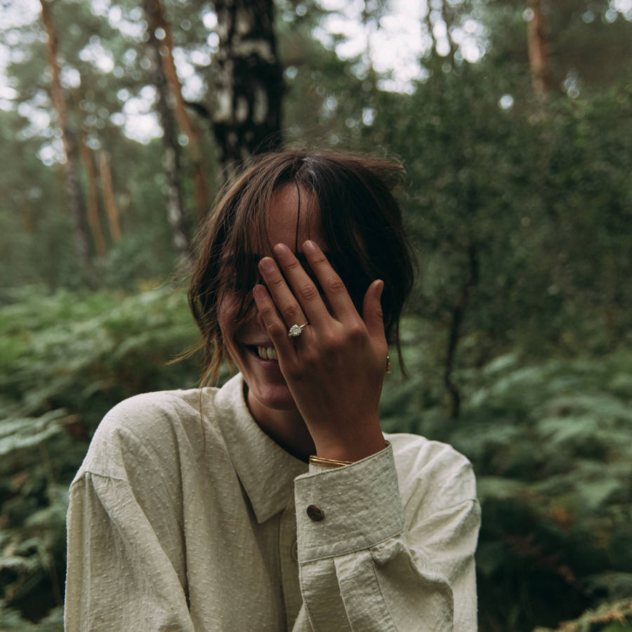 Woman smiling in a forest, covering part of her face while showing a bespoke diamond engagement ring on her hand.