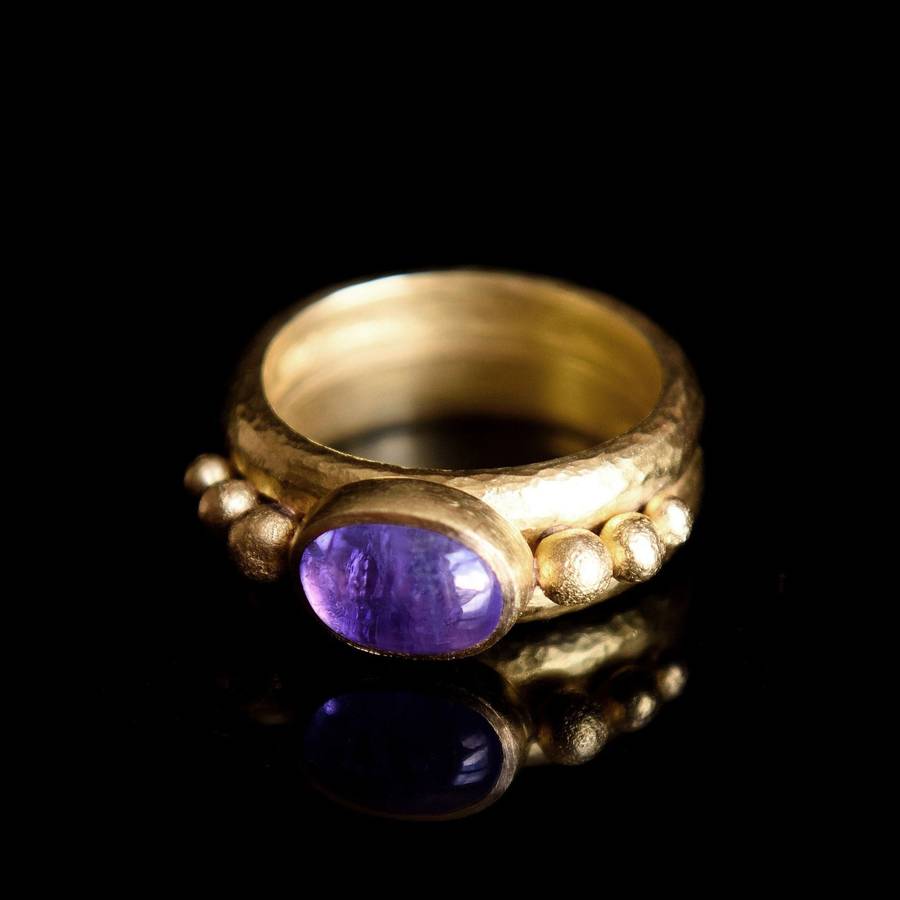 Gold ring with an oval purple tanzanite cabochon set in a bezel, flanked by small textured gold beads on a hammered band, photographed on a black reflective surface