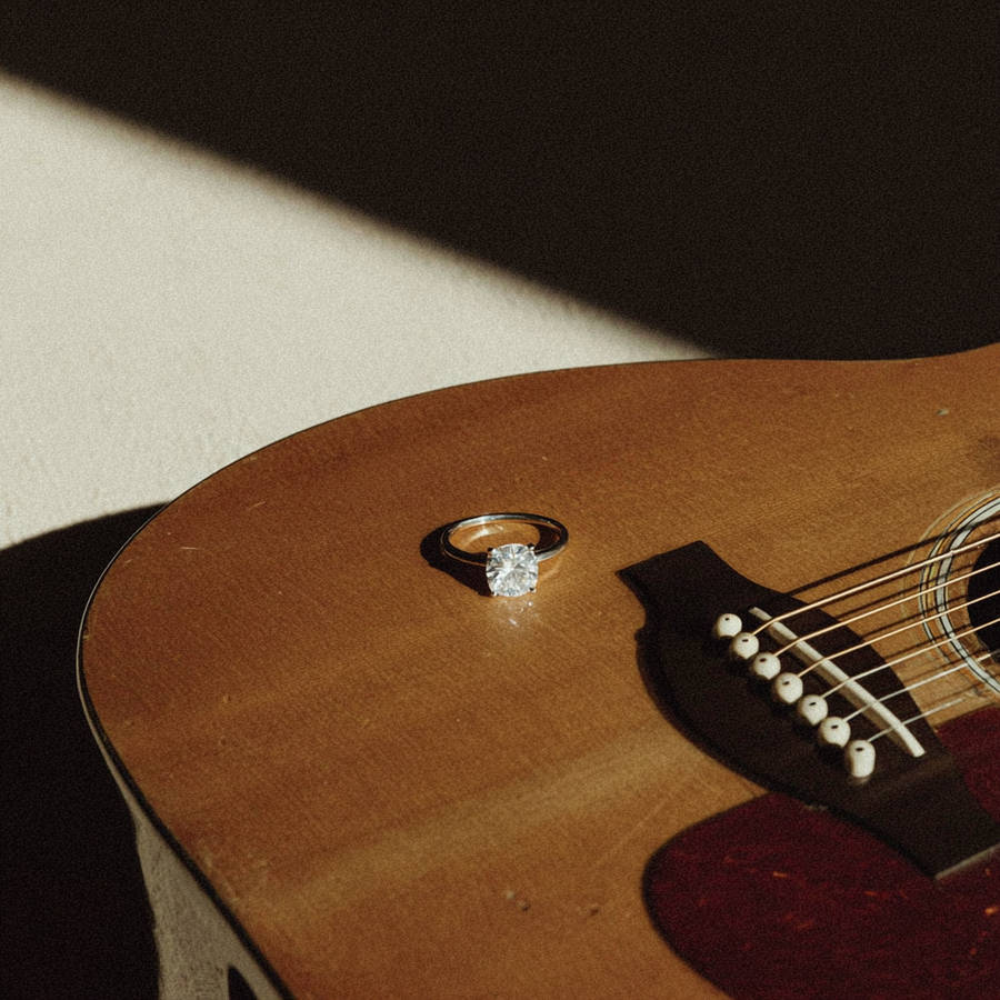 Solitaire diamond engagement ring resting on an acoustic guitar, illuminated by warm natural light and soft shadows.