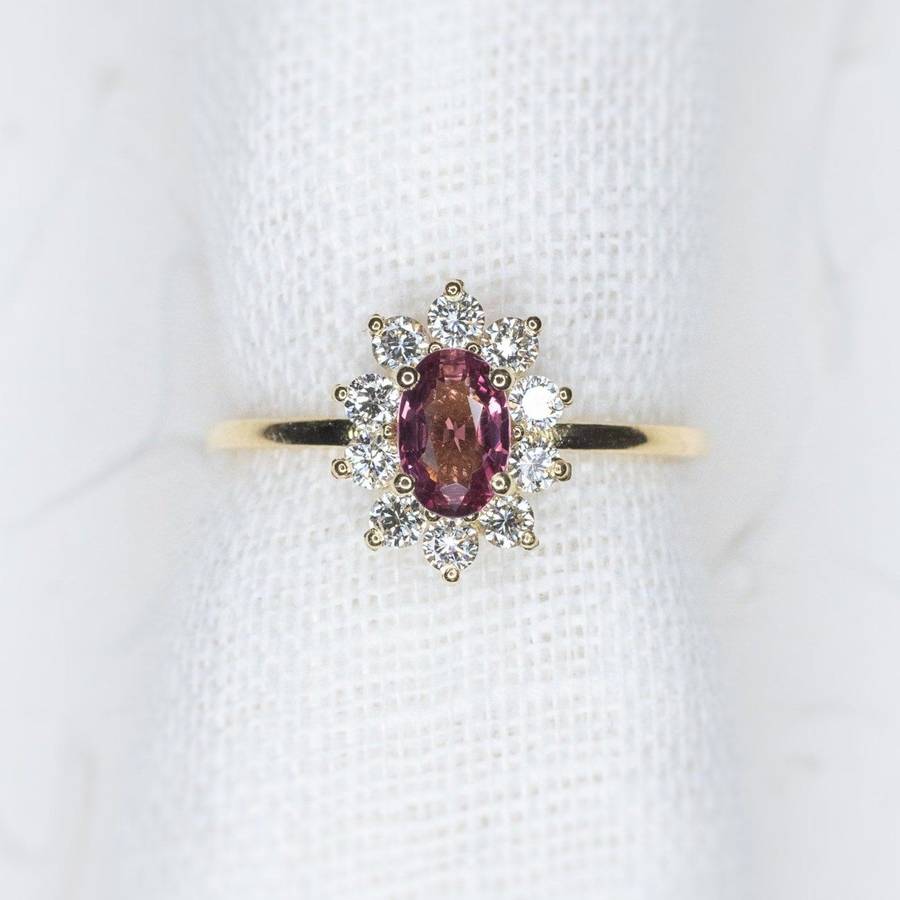 Oval pink sapphire halo ring with a diamond surround on a slim yellow gold band, photographed against a soft white textured background