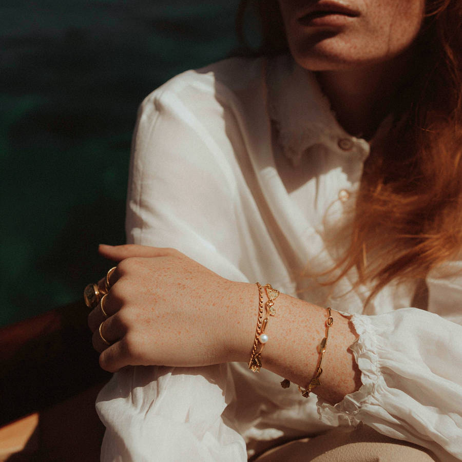Close-up of a woman’s freckled wrist and hand resting across her body, wearing layered delicate gold bracelets, dressed in a soft white blouse, lit by warm natural light.