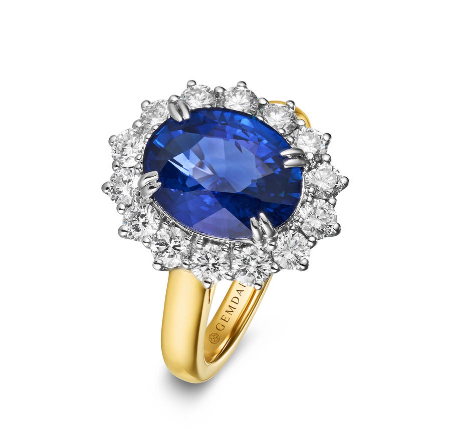 Oval blue sapphire ring with a halo of round diamonds, set in a yellow gold band with white metal prongs, photographed on a white background.