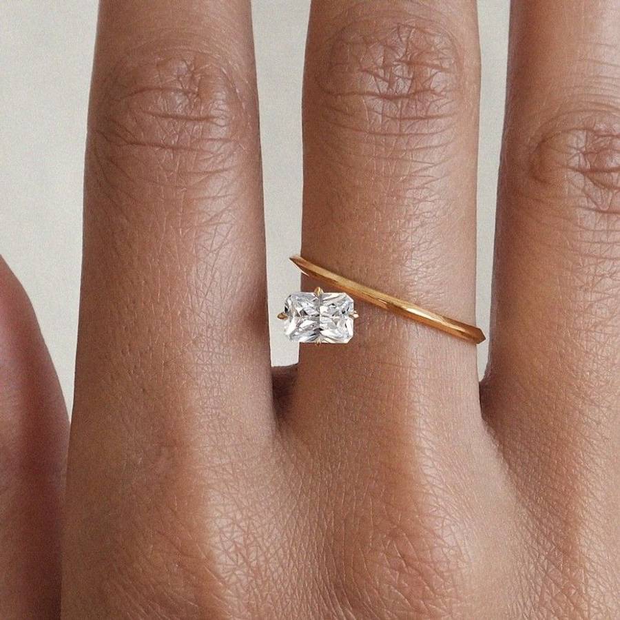 Hand wearing a minimalist yellow gold ring with a horizontally set emerald-cut diamond and a slender angled gold band crossing above the stone.