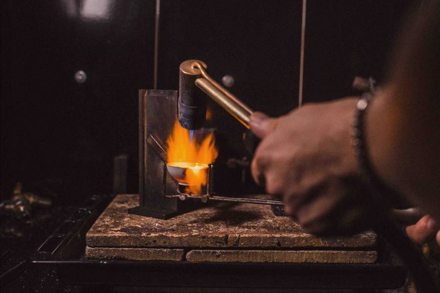 Jeweller uses a torch and crucible to melt metal in a workshop, flames glowing over a firebrick workbench.