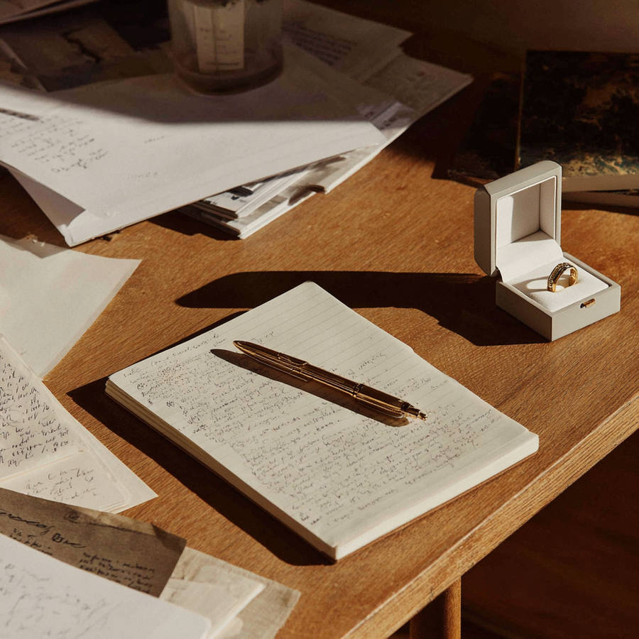 Open ring box with bespoke gold engagement ring beside handwritten notes and pen on sunlit desk