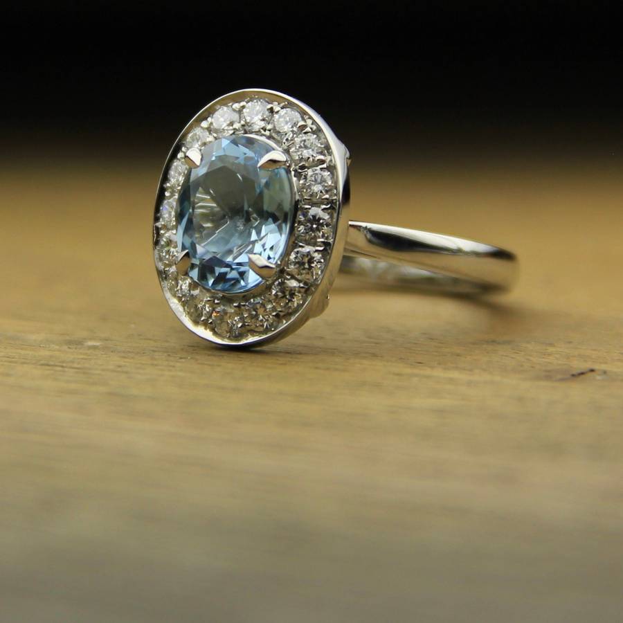 White gold ring with an oval light blue aquamarine center stone surrounded by a halo of small diamonds, resting on a wooden surface.