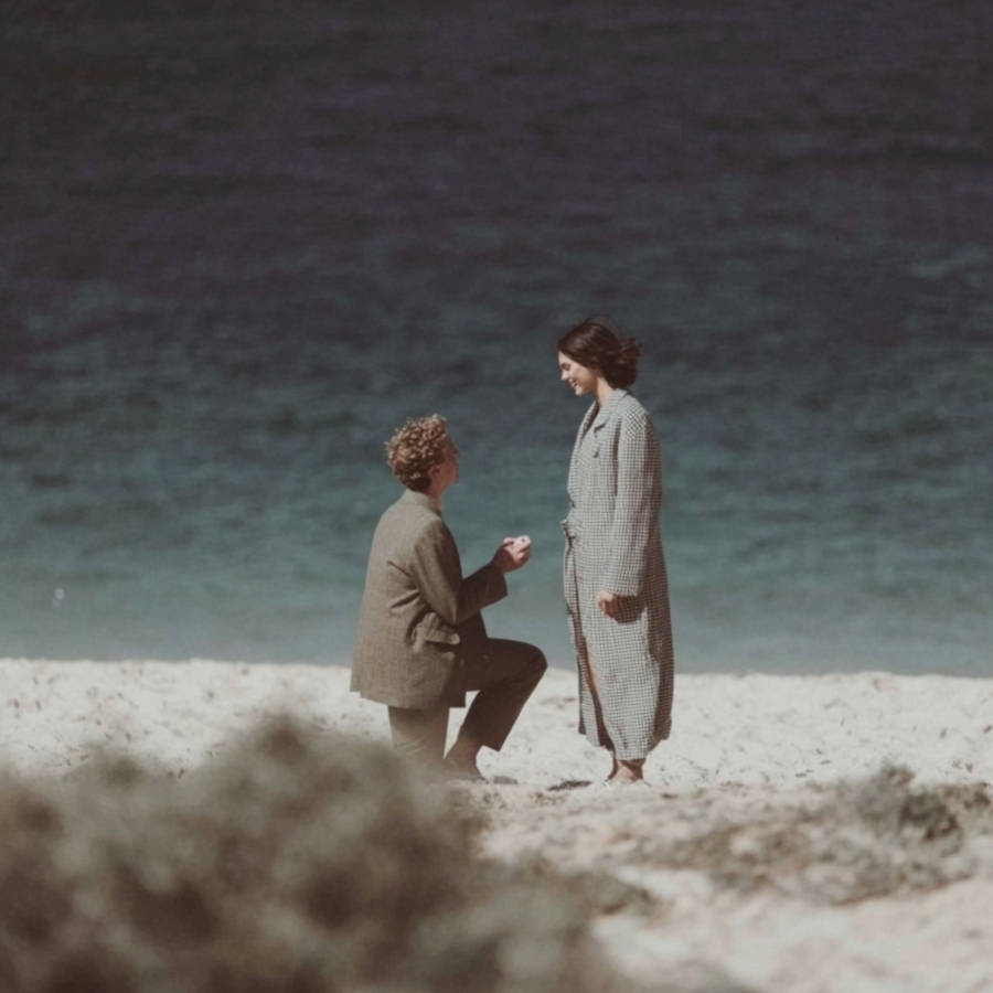 Man kneels proposing with a ring box to a woman on a sandy beach, ocean backdrop, vintage-style photo