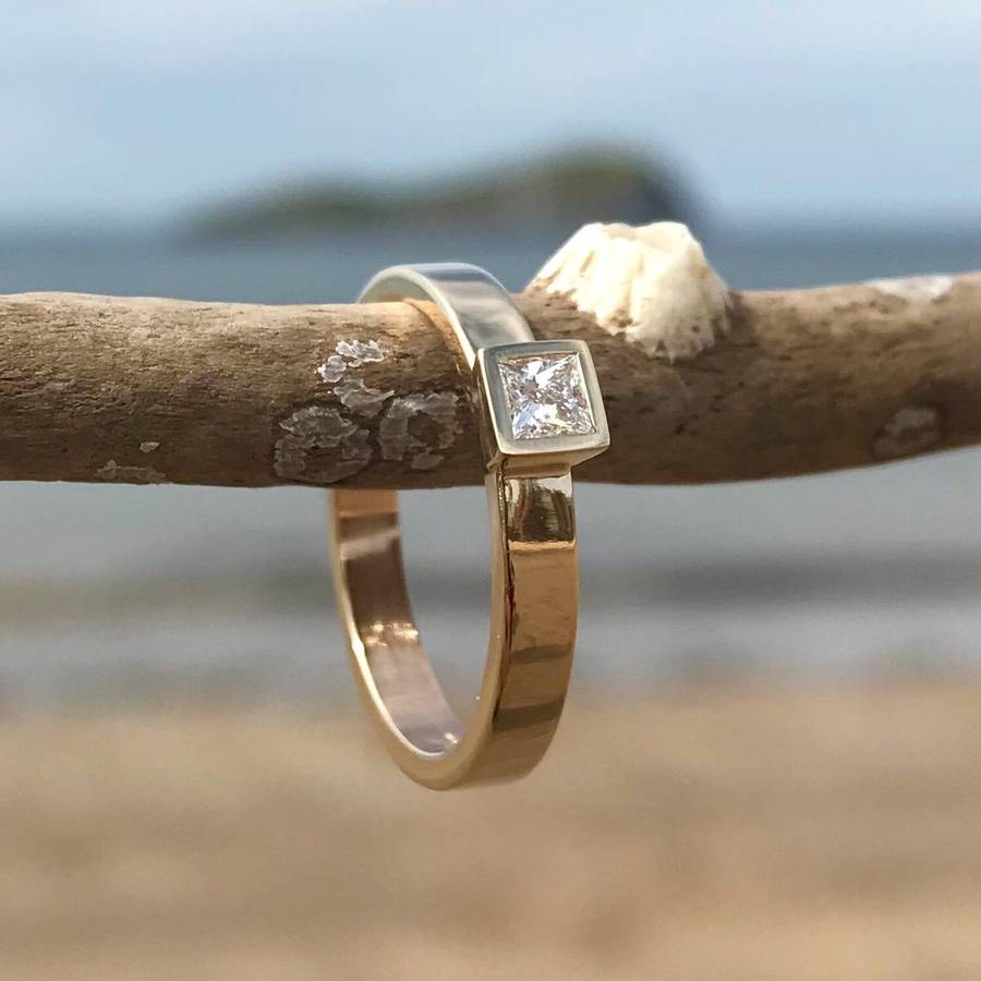 Two-tone gold ring with a square bezel-set diamond hanging from a weathered branch, photographed outdoors with a softly blurred beach and sea in the background.
