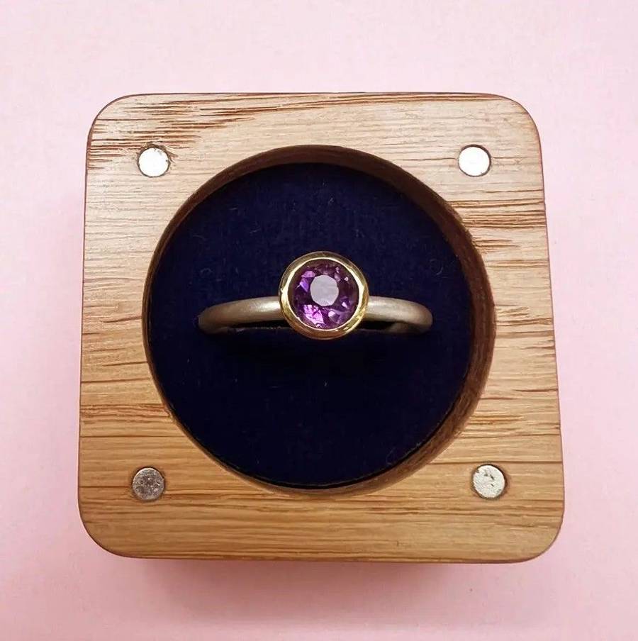 Custom 18ct gold ring with bezel-set amethyst, displayed in a wooden box on a pink background
