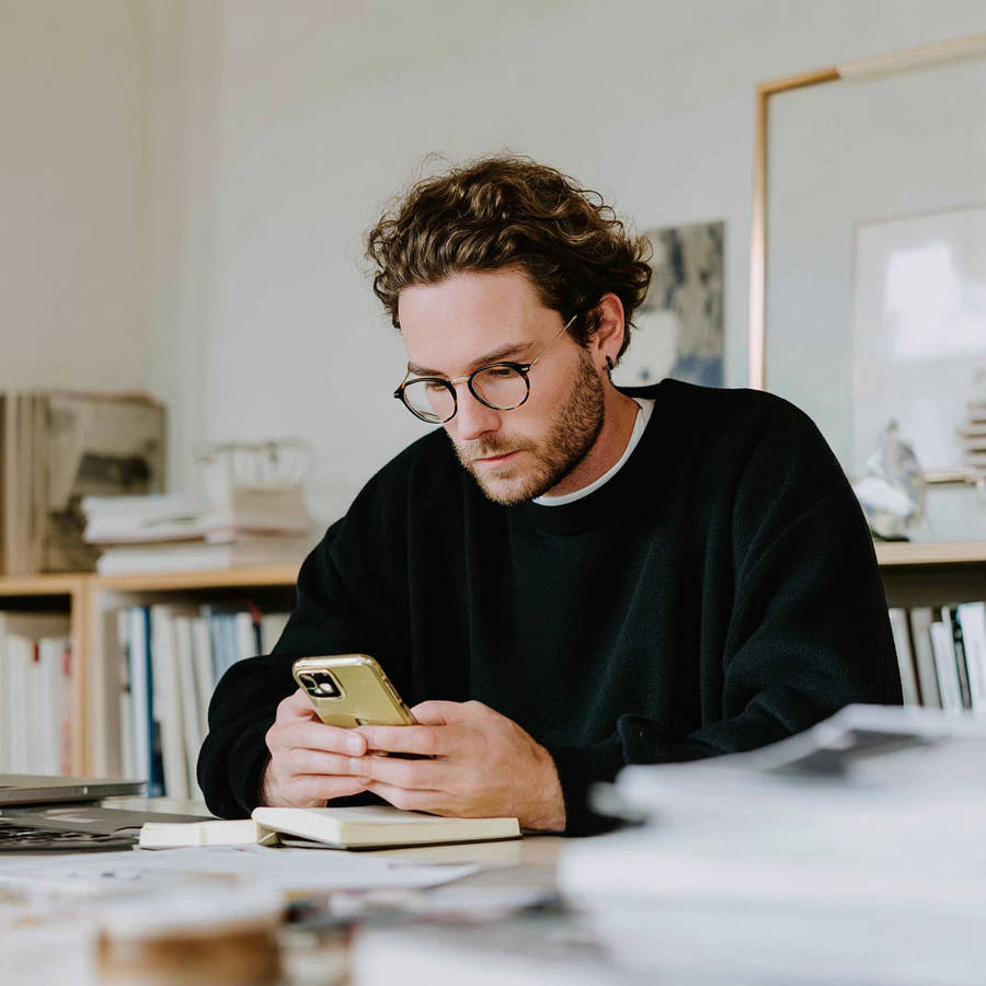 Man wearing glasses looking at smartphone while sitting at desk with notebook and papers in a bright home office