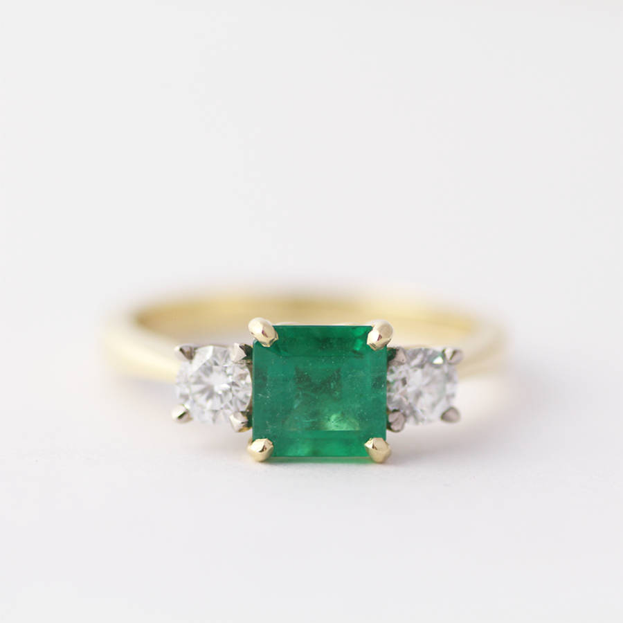 Emerald-cut green emerald flanked by two round white diamonds set in a yellow gold band against a white background