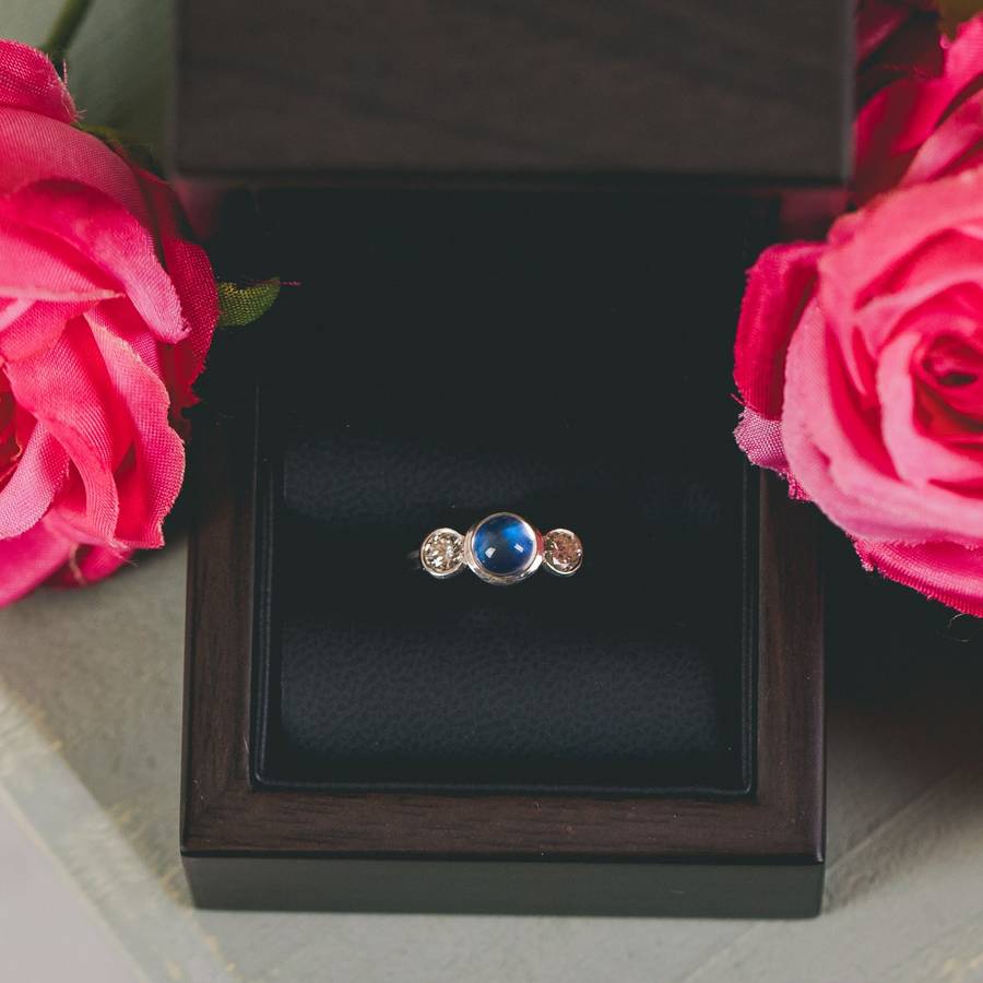 Three-stone ring with a central blue moonstone and two smaller clear gemstones, displayed inside a dark wooden ring box, framed by bright pink roses on either side.