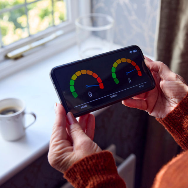 Senior Woman With Smart Meter Trying To Keep Warm By Radiator During Cost Of Living Energy Crisis - How Can We Support Households with Energy Bills