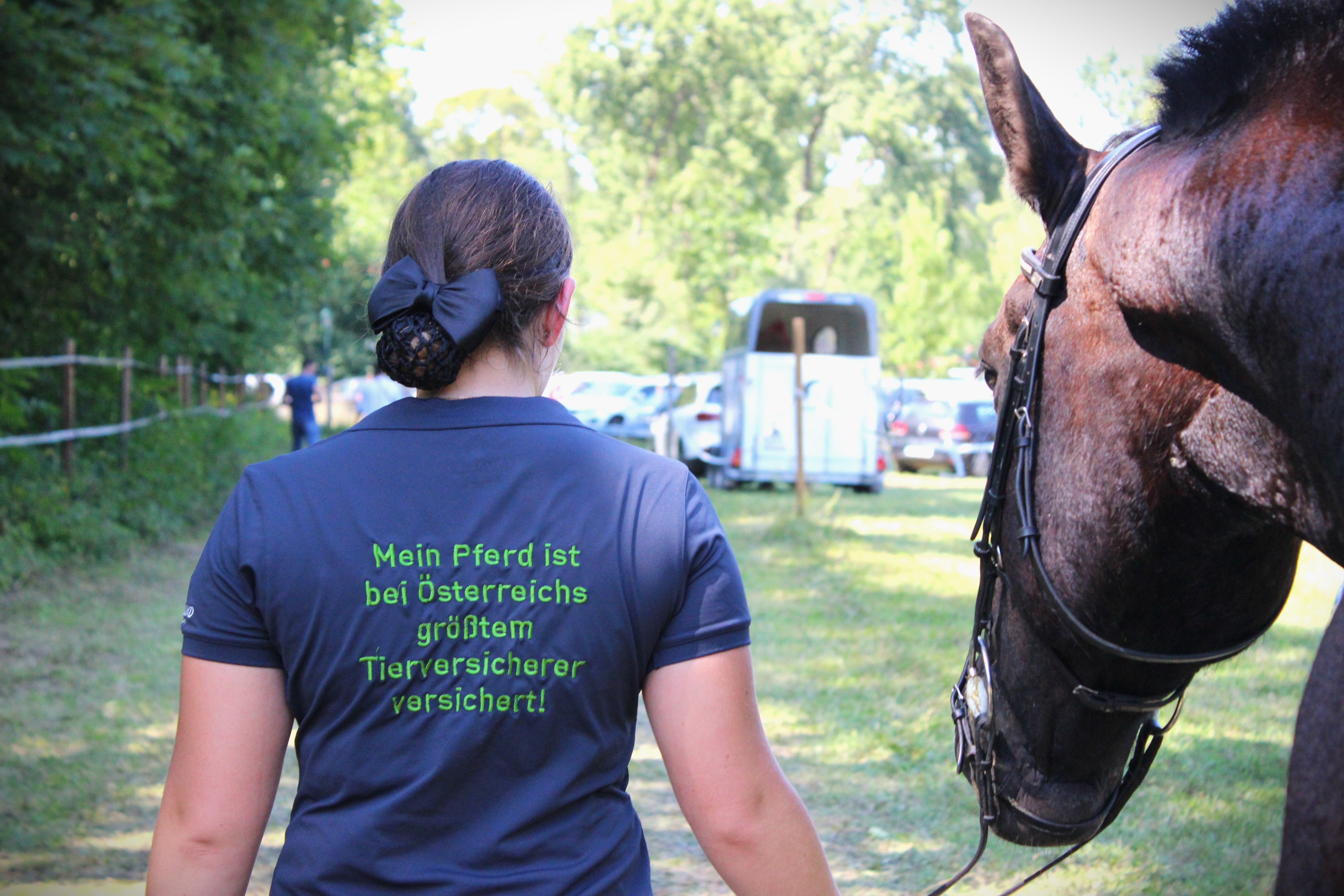 Rückenansicht einer Frau, die ein Pferd am Strick führt und auf ihrem T-Shirt die Aufschrift "Mein Pferd ist bei Österreichs größtem Tierversicherer versichert!" trägt.