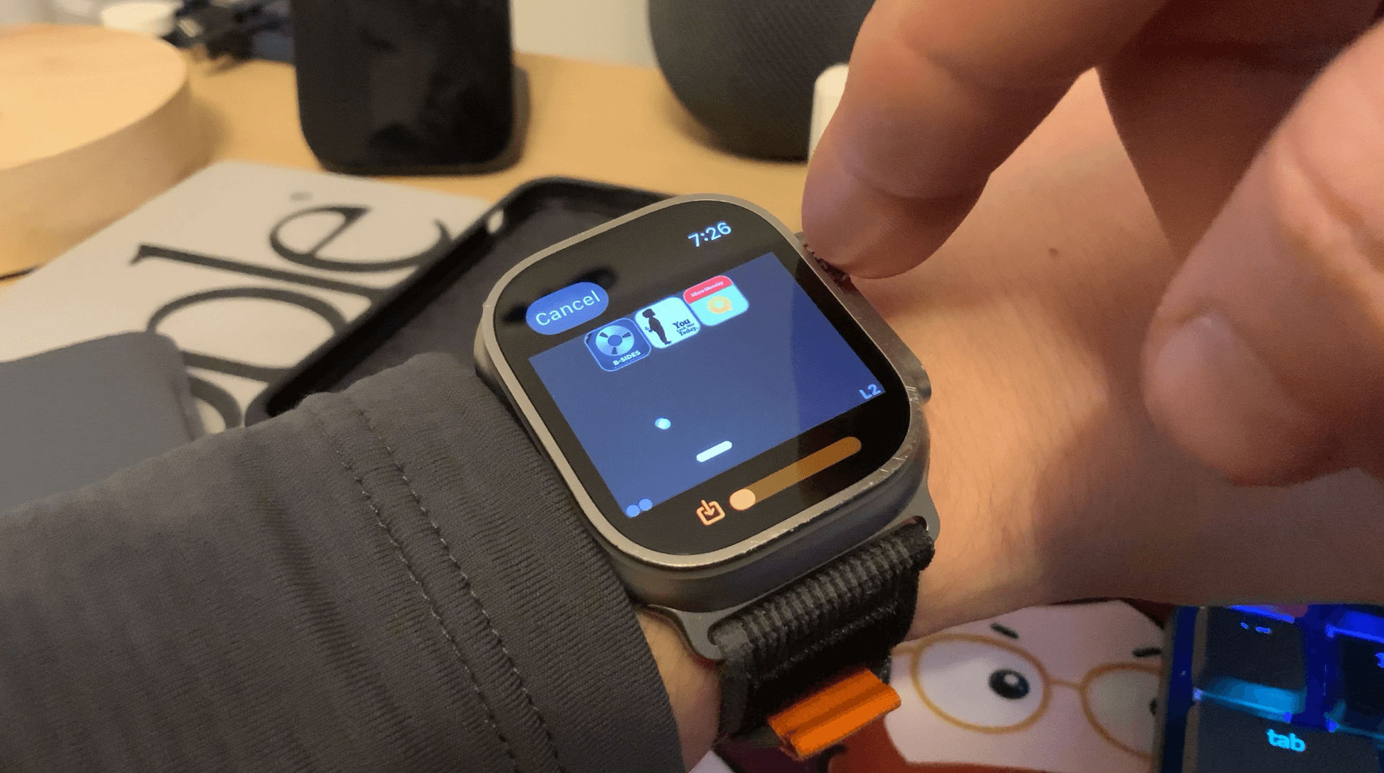 A smartwatch on a wrist displays a game of Breakout with podcast artwork as the tiles; a finger prepares to tap the screen. A blurred office environment with various items forms the background.