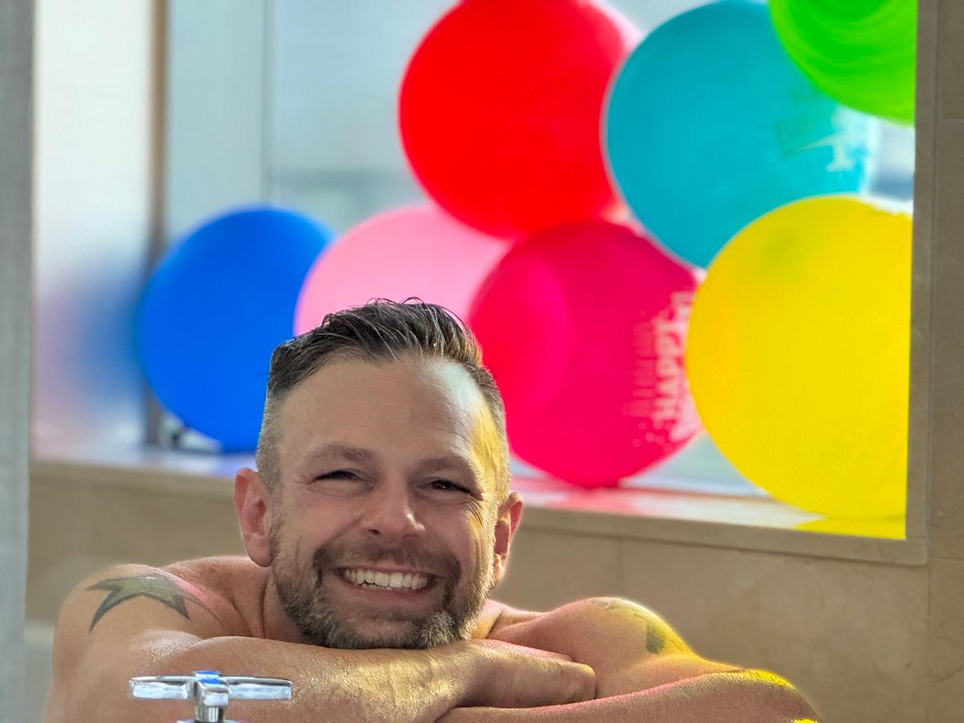 Joe smiling with a wall of colorful balloons behind him on his birthday