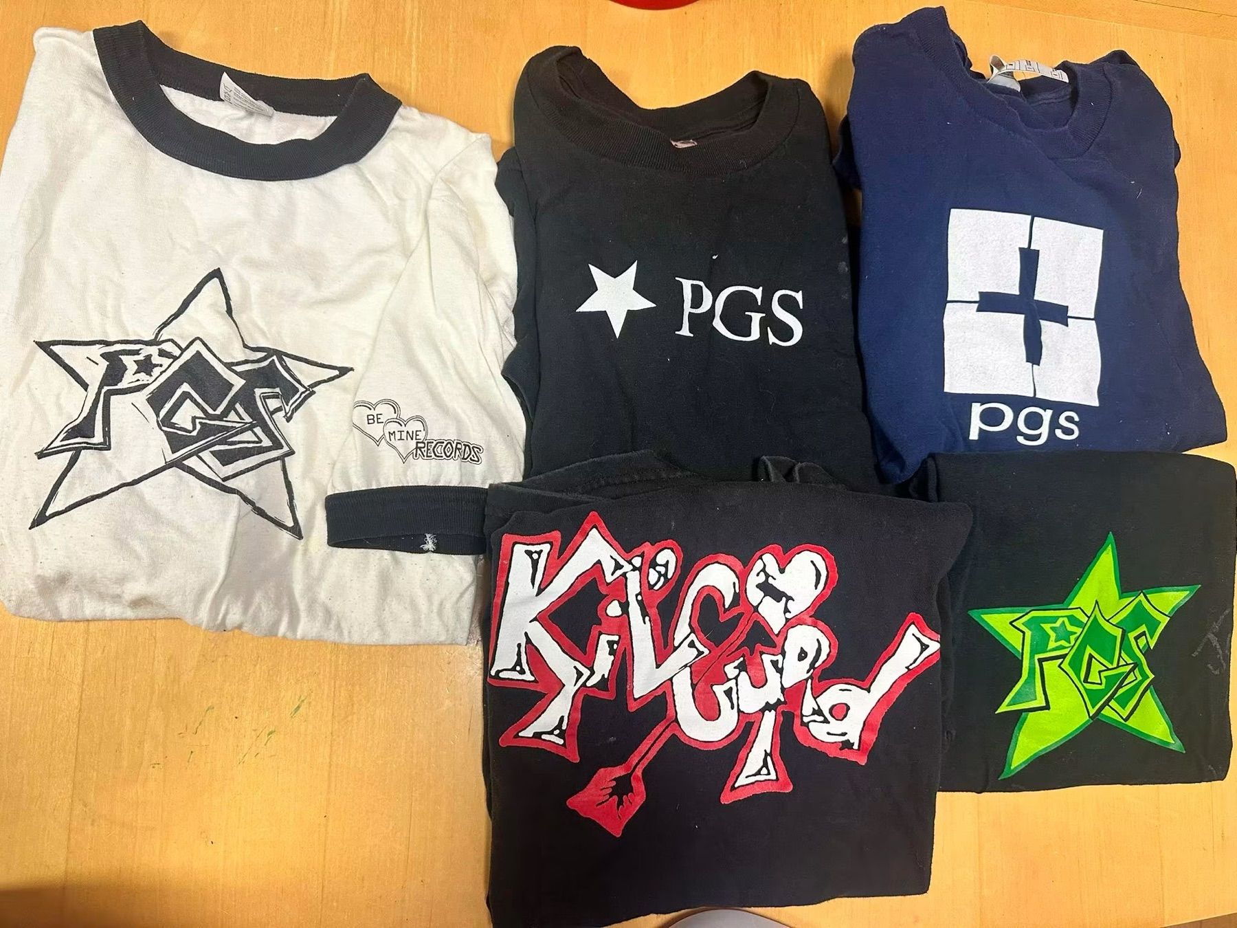 Five vintage PGS band t-shirts with hand-drawn logos, laid out on a table