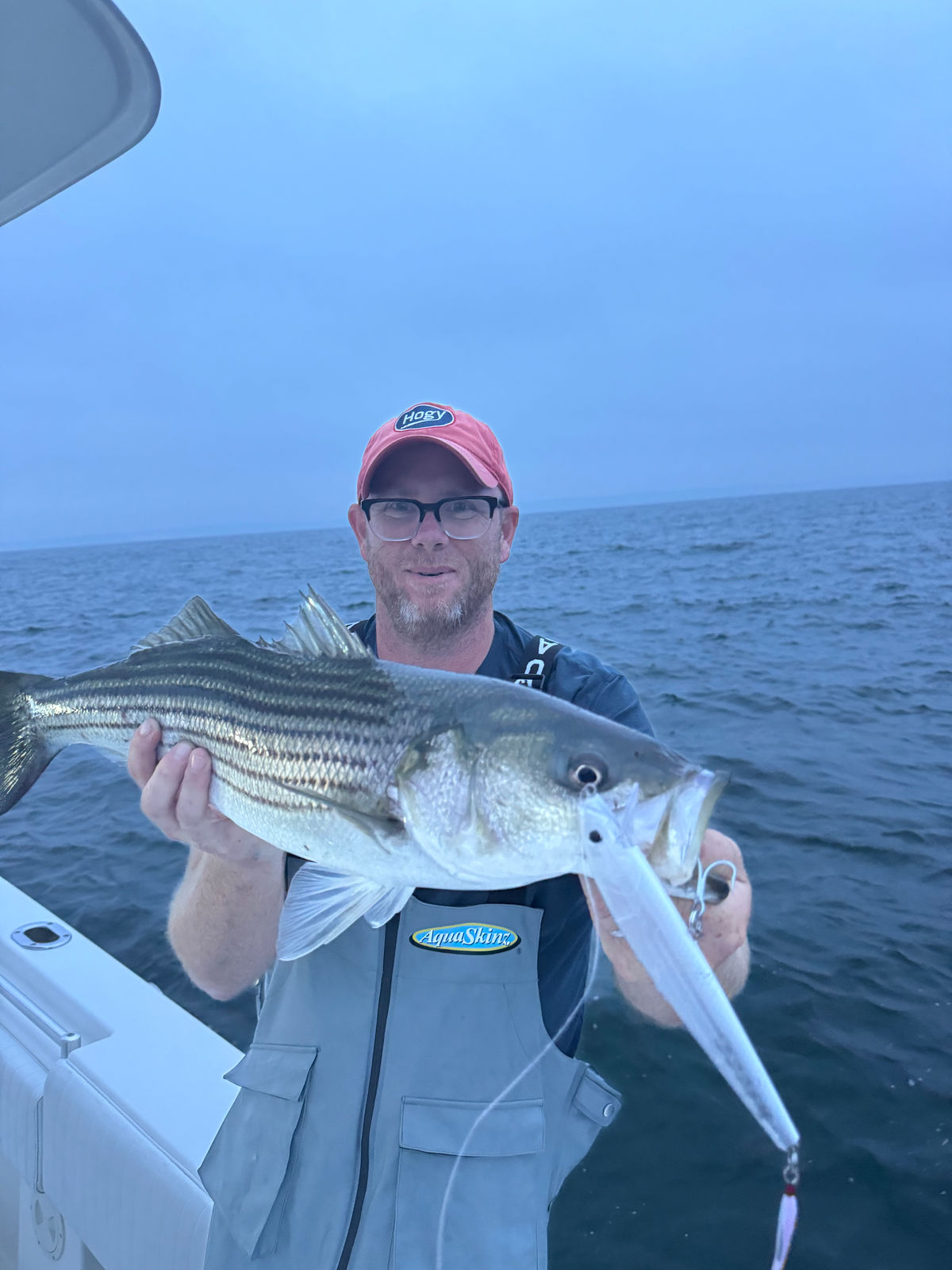 A striped bass held horizontally aboard the boat with an OG Swimmer clearly visible in its mouth.