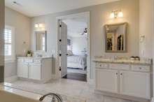 This dreamy primary suite bath includes separate vanities flanking the door with white cabinetry and natural stone counters