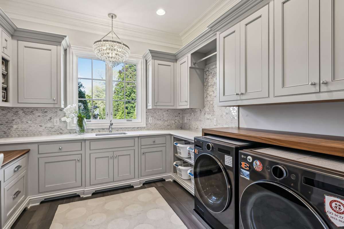 Plank tile flooring is complemented by a full-height leaf patterned marble backsplash. Practical features include laundry basket storage, and a hanging rod. A sparkling crystal chandelier and an LG washer and dryer complete this space.
