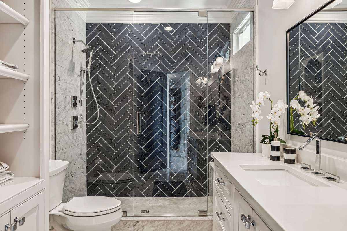 The ensuite bath features a walk-in shower with a herringbone glass tile wall enclosed by a frameless glass surround with a transom window and bench. Additional features include a vanity with quartz, Carrara marble tile flooring, and linen storage.