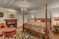 View of the master bedroom suite, featuring hardwood flooring, an electric fireplace, charming wall and ceiling paneling constructed of reclaimed wood, and and elegant chandelier with graceful medallion mounting.