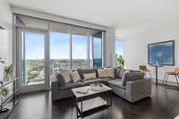 Floor-to-ceiling windows illuminate the living room with stunning skyline views, while motorized shades provide privacy and gentle light diffusion even on the sunniest days. The herringbone wood floors add a touch of elegance and beauty to the space.