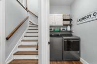 A roomy laundry nook is prepared to accommodate your washer, dryer, and key housekeeping essentials with wall-to-wall shelving and convenient cabinet space at the base of the staircase offering convenience and functionality to your household routine