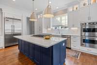 Sub zero fridge and large island with blue cabinetry. Beautiful light fixtures over the island and extra lighting in high cabinetry