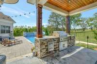 Outdoor living area with a covered kitchen, built-in grill, stone accents, and poolside views, perfect for entertaining.