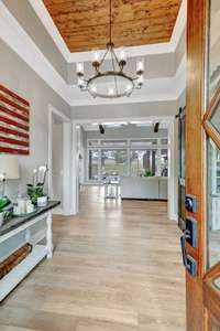 Inviting foyer with wooden ceiling, elegant chandelier, and open view into a bright living space with large windows.