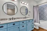 A stylish double vanity guest bathroom featuring elegant granite countertops and round decorative vanity mirrors. The space includes a tiled surround tub and shower combo, offering both functionality and charm.