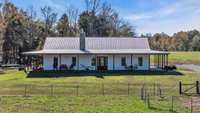 A pristine white farmhouse built in 2021, featuring clean lines, metal roof, abundance of windows and a welcoming 2500 square ft wraparound porch with the most beautiful views.