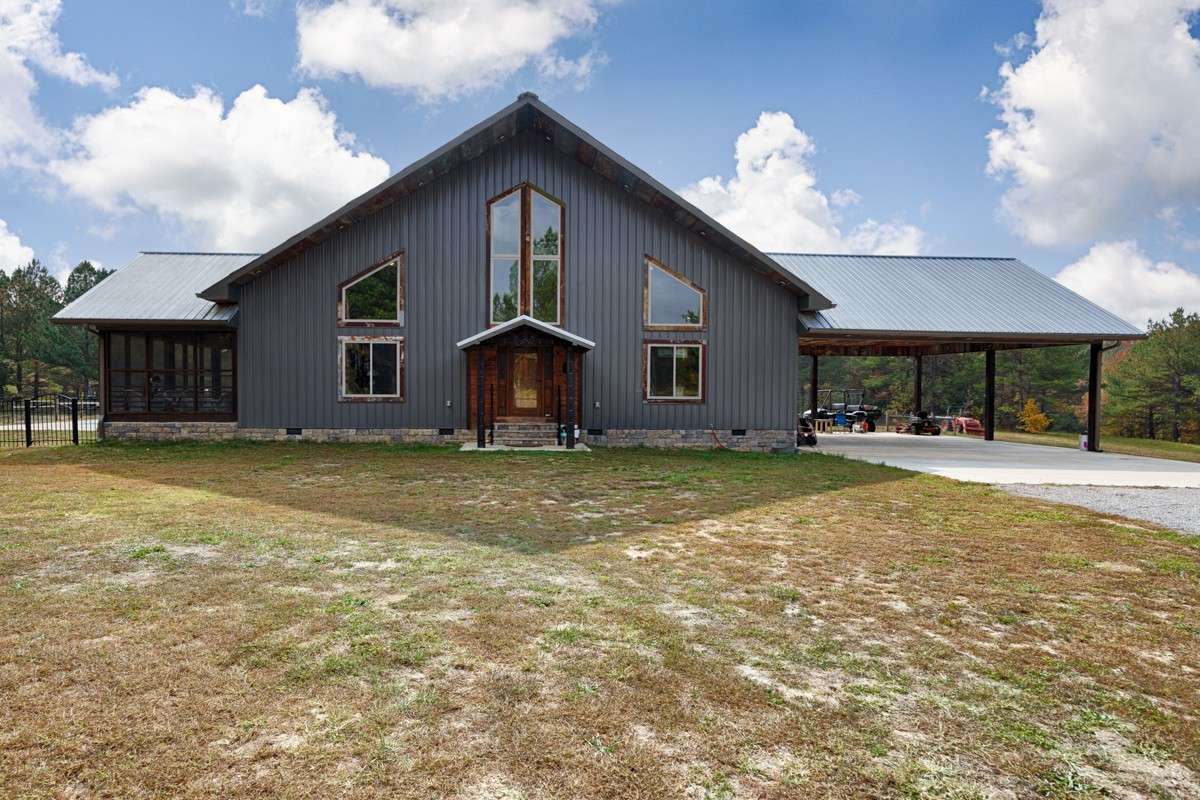 Front of house- gravel drive, concrete pad for parking and carport, metal building and roofing, large windows, covered front entry, screened porch