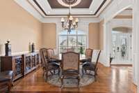The formal dining room has a custom ceiling design above the chandelier as well as ornate molding, gorgeous hardwood flooring, and plantation shutters on the large windows.